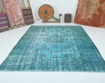 May include: A large, rectangular turquoise rug with a distressed, vintage appearance. The rug features a central medallion design and a border with geometric patterns. Other rugs and decorative items are in the background.