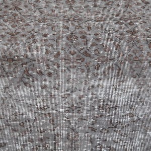 May include: Close-up of a gray rug with a distressed, vintage appearance. The rug features a repeating pattern of brown floral motifs against a gray background. The design has a faded, textured look, suggesting it is an antique or a rug with a worn aesthetic.