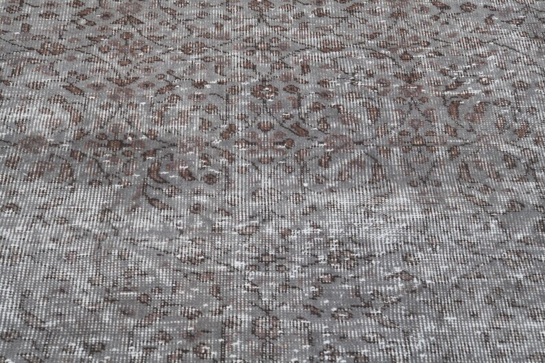 May include: Close-up of a gray rug with a distressed, vintage appearance. The rug features a repeating pattern of brown floral motifs against a gray background. The design has a faded, textured look, suggesting it is an antique or a rug with a worn aesthetic.
