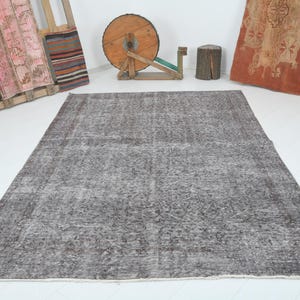 May include: A large, rectangular, gray rug with a faded, patterned design. The rug is the focal point, set against a white background. Various decorative items are in the background, including a wooden spool and a wooden pallet with colorful textiles.