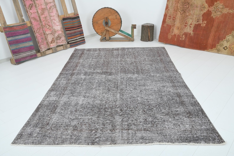 May include: A large, rectangular, gray rug with a faded, patterned design. The rug is the focal point, set against a white background. Various decorative items are in the background, including a wooden spool and a wooden pallet with colorful textiles.