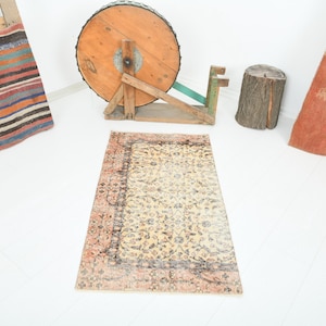 May include: A rectangular, vintage-style rug with a central floral pattern in cream and beige, framed by a faded reddish-brown border. The rug is placed on a white surface, with other decorative items in the background.