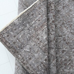 May include: A section of a gray vintage rug with a distressed, floral pattern. The rug has a faded appearance with brown and white accents. The edge of the rug is visible, showing a cream-colored fringe.
