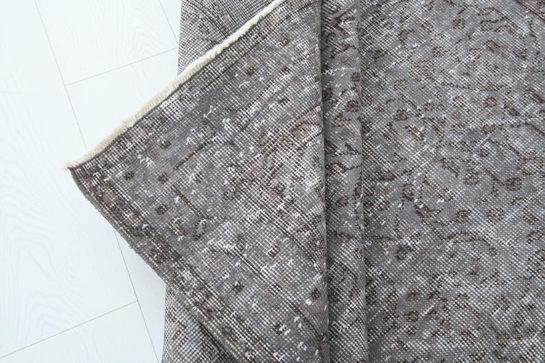 May include: A section of a gray vintage rug with a distressed, floral pattern. The rug has a faded appearance with brown and white accents. The edge of the rug is visible, showing a cream-colored fringe.