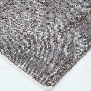 May include: Close-up of a distressed, vintage-style rug with a faded, floral pattern. The rug features a palette of gray, brown, and cream colors. The edges are frayed, adding to the rug's antique aesthetic. The rug is placed on a white surface.