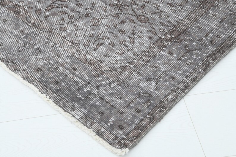 May include: Close-up of a distressed, vintage-style rug with a faded, floral pattern. The rug features a palette of gray, brown, and cream colors. The edges are frayed, adding to the rug's antique aesthetic. The rug is placed on a white surface.