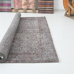 May include: A rolled-up, rectangular, gray rug with a floral pattern. The rug is partially unrolled, revealing its intricate design and color variations. The rug is placed on a white surface.