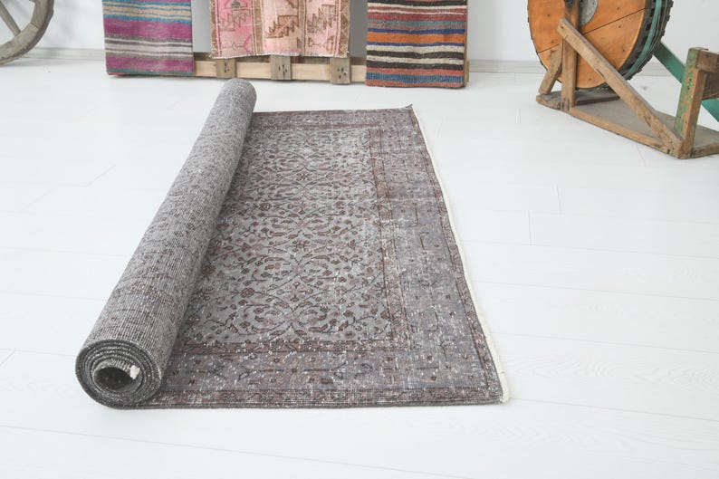 May include: A rolled-up, rectangular, gray rug with a floral pattern. The rug is partially unrolled, revealing its intricate design and color variations. The rug is placed on a white surface.