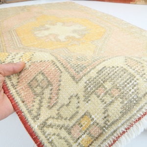 May include: A vintage Turkish rug with a cream background and a central medallion in shades of yellow and pink. The rug has a red border with fringe.