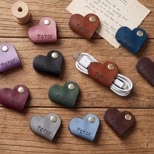 Puede incluir: Una colección de organizadores de cables de cuero en forma de corazón en varios colores, incluyendo marrón, azul y morado. Cada organizador está personalizado con el nombre "Peter" y asegurado con un broche metálico. Un cable blanco está envuelto.