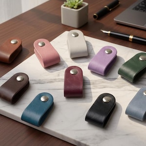 Puede incluir: Una colección de organizadores de cables de cuero en varios colores, incluyendo marrón, rosa, gris, morado, verde, burdeos, turquesa, negro y azul. Cada organizador presenta un diseño curvo con un cierre de botón metálico. Los organizadores se exhiben sobre una superficie de mármol.