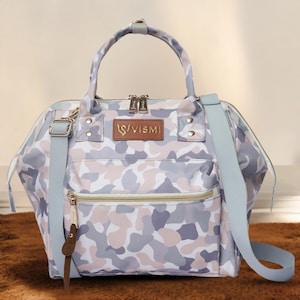 Könnte beinhalten: Handtasche mit Camouflage-Muster, mit einem oberen Griff und einem abnehmbaren Schultergurt. Die Tasche hat eine Reißverschlusstasche vorne, ein braunes Lederetikett und einen Logoaufnäher mit der Aufschrift "VISMI". Die Farbpalette umfasst Grau, Rosa und Weiß.