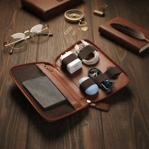 以下が含まれることがあります： 茶色の革製トラベルオーガナイザー。テクノロジーアクセサリーを表示するために開いています。内部には、白い充電器、ケーブル、茶色のストラップで固定されたイヤホンがあります。小さな青い丸い物体も固定されています。