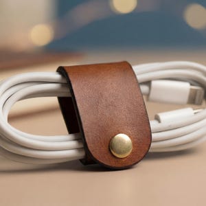 Puede incluir: Un organizador de cables de cuero marrón con un cierre de botón dorado sujeta un cable de carga blanco. El cable está cuidadosamente enrollado, mostrando un diseño minimalista. El organizador es de cuero marrón.