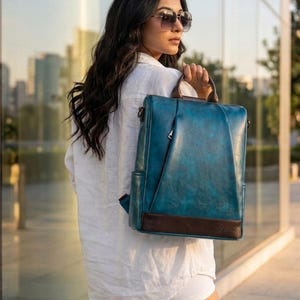 Peut inclure: Un sac à dos rectangulaire bleu sarcelle avec une finition vieillie, doté d'une base en similicuir marron foncé, d'une poignée supérieure et d'une poche frontale zippée verticale. Le sac possède des poches latérales et des bretelles réglables.