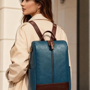 Puede incluir: Una mochila rectangular de color azul cerceta con ribetes de piel sintética marrón oscuro, correas ajustables, un asa superior y un bolsillo frontal vertical con cremallera. La base tiene un panel marrón oscuro con un logotipo grabado.