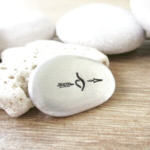 May include: A small, silver-coloured, oval-shaped stone with an arrow and a swirl design engraved on it.