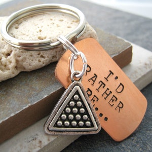 May include: A copper keychain with the text "I'd Rather Be..." and a silver triangle charm with a pool ball rack design.