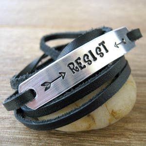 May include: Black leather wrap bracelet with a silver metal tag that says "Resist" with an arrow pointing to the right.