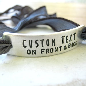 May include: A silver bracelet with a gray ribbon. The bracelet has a metal tag that says "CUSTOM TEXT ON FRONT & BACK".
