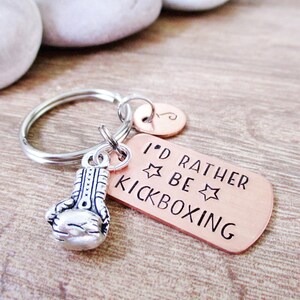 May include: A copper-colored keychain with a silver boxing glove charm. The keychain has the text "I'd Rather Be Kickboxing" with two stars.