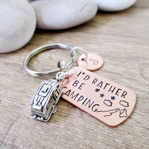 May include: A copper-colored keychain with the text "I'd rather be camping" and a small silver camper charm. The keychain also has a small silver circle with the letter "J" on it.