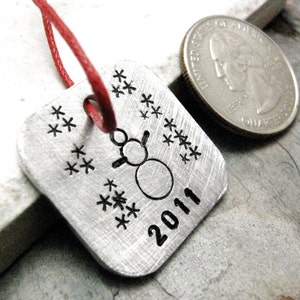 May include: Silver metal ornament with a snowman design and the year 2011. The ornament is decorated with snowflakes and has a red string attached.