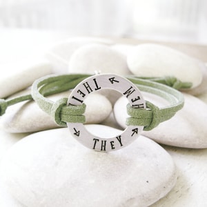 May include: A green suede bracelet with a silver circular charm that reads "They Them" in a circular font.