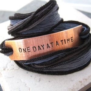 May include: A copper bracelet with the words "One Day At A Time" engraved on it. The bracelet is wrapped in black and gray ribbon.