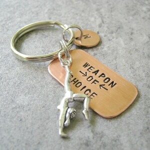 May include: A copper keychain with a silver gymnast charm and the text "Weapon of Choice".