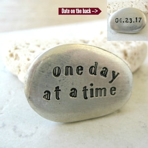 May include: A silver-toned metal rock with the phrase "one day at a time" engraved on it. The back of the rock has the date "04.23.17" engraved on it.