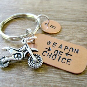 May include: A silver motorcycle charm attached to a copper keychain with the words "Weapon of Choice" engraved on the tag. The keychain also has a small copper charm with the letter "H" on it.