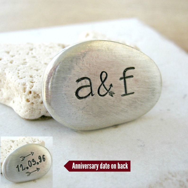 May include: A silver metal charm with the initials "a&f" engraved on the front. The back of the charm has an anniversary date engraved with arrows pointing to the date "12.03.96".