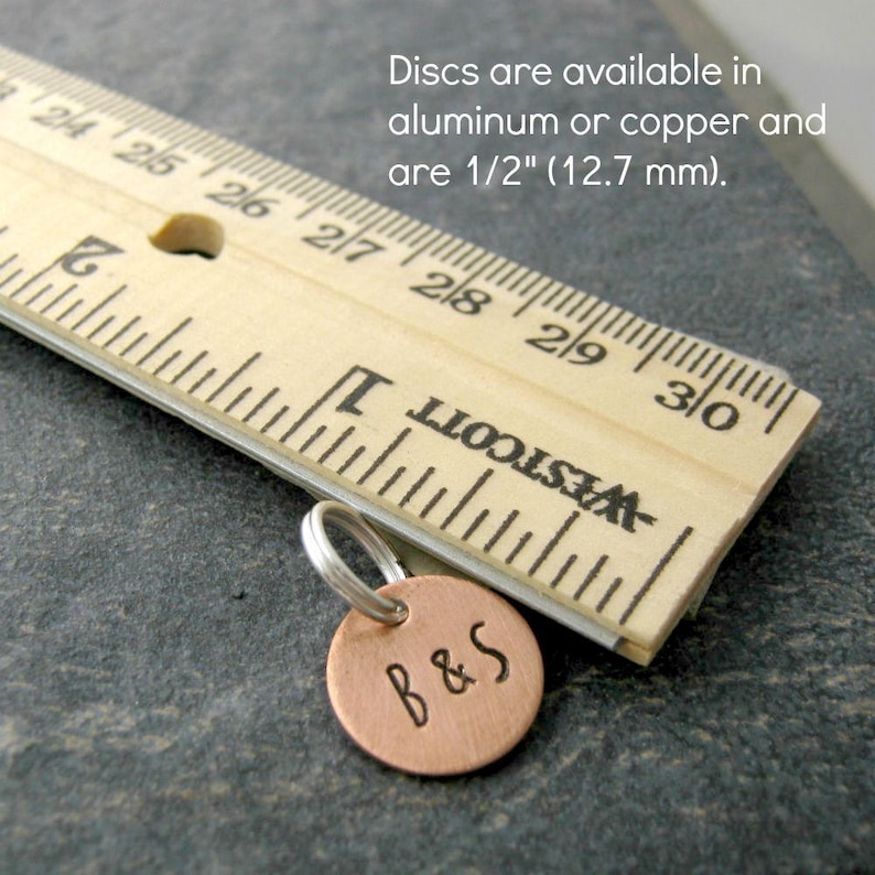 May include: A close-up of a copper disc charm with the initials "B&S" engraved on it. The charm is 1/2 inch (12.7 mm) in diameter and is attached to a silver jump ring. The charm is sitting on a wooden ruler.