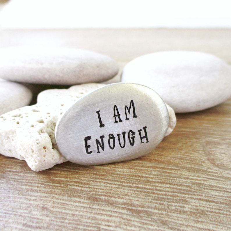 May include: A silver metal token with the words "I AM ENOUGH" engraved on it. The token is resting on a white rock.
