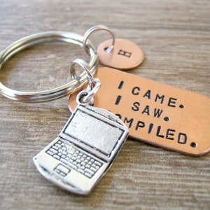 May include: A silver-colored laptop charm keychain with a copper tag that reads "I CAME. I SAW. COMPILED." The keychain also includes a small, round copper tag with the letter "B". The keychain is attached to a silver key ring.