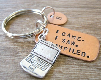 May include: A silver-colored laptop charm keychain with a copper tag that reads "I CAME. I SAW. COMPILED." The keychain also includes a small, round copper tag with the letter "B". The keychain is attached to a silver key ring.