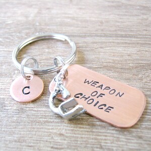 May include: A copper keychain with a silver charm and a round copper tag with the letter "C" engraved on it. The tag reads "Weapon of Choice".