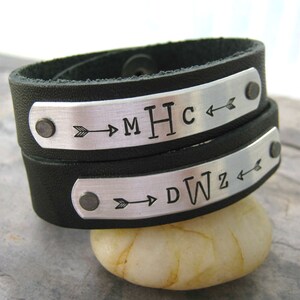 May include: Two black leather bracelets with silver metal plates. The plates have arrows and the initials "MHC" and "DWZ" engraved on them.