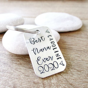 May include: Silver keychain with the text "Best Nana Ever 2020" and "Caroline" written vertically on the side. The keychain has a heart symbol at the top and bottom.