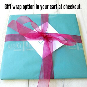 K&ouml;nnte beinhalten: Eine t&uuml;rkisfarbene Geschenkbox mit einem rosa Band, das zu einer Schleife gebunden ist. Auf der Box steht "Risky Be" gedruckt.