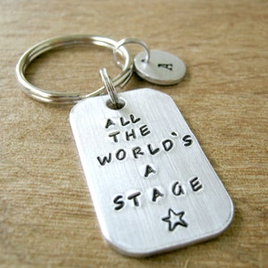 May include: Silver keychain with a tag that reads "All the world's a stage" and a small star. The keychain also has a silver charm with the letter "A" on it.