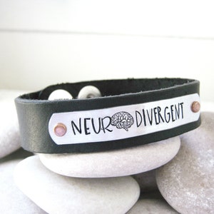 May include: Black leather bracelet with a silver plate engraved with the text "Neurodivergent" and a brain symbol.