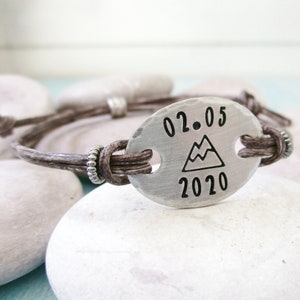 May include: A brown leather bracelet with a silver oval charm. The charm has the date "02.05.2020" and a mountain icon engraved on it.
