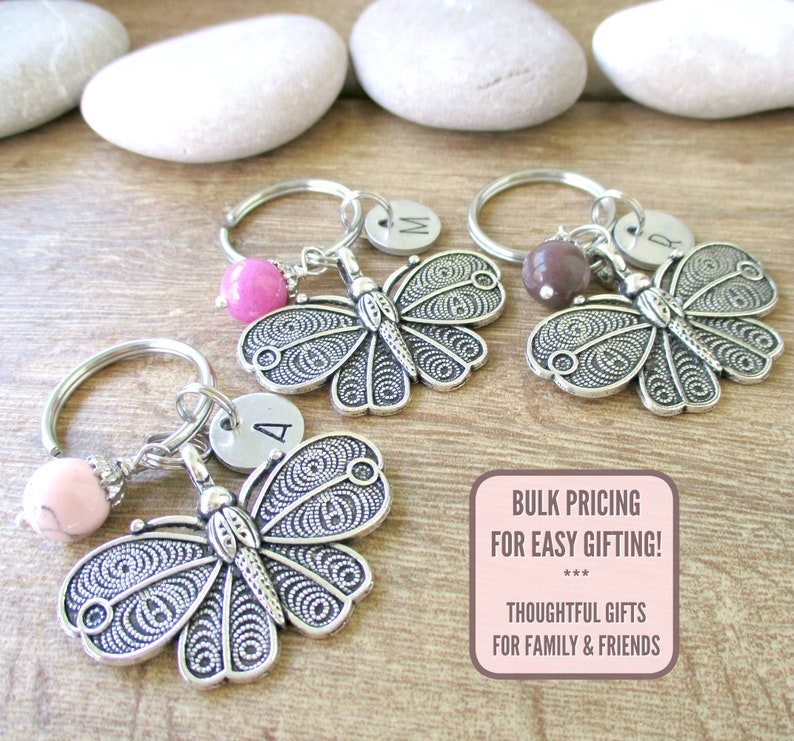 Puede incluir: Tres llaveros de mariposa plateados con un peque&ntilde;o dije y una inicial de letra en un disco plateado. Los dijes son rosa, marr&oacute;n y rosa claro. Los llaveros est&aacute;n sobre una superficie de madera.