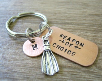 May include: A copper keychain with a silver flipper charm and a copper tag that reads "Weapon of Choice" with arrows pointing to the word "Choice". The keychain also has a small copper disc with the letter "M" engraved on it.
