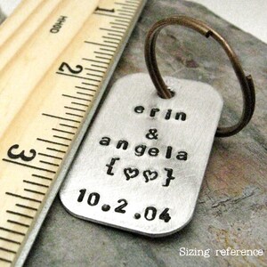 May include: Silver keychain with the text "erin & angela [heart] 10.2.04" engraved on it. The keychain is next to a wooden ruler for size reference.