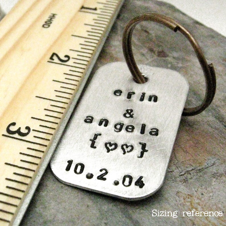 May include: Silver keychain with the text "erin & angela [heart] 10.2.04" engraved on it. The keychain is next to a wooden ruler for size reference.