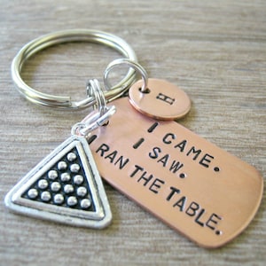 May include: A copper keychain with a silver triangle charm featuring a pool ball rack design. The keychain has a tag that reads "I came. I saw. I ran the table."