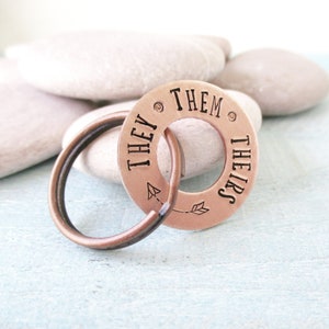 May include: Copper keyring with a round tag that says "They/Them/Theirs" with an arrow on each side.